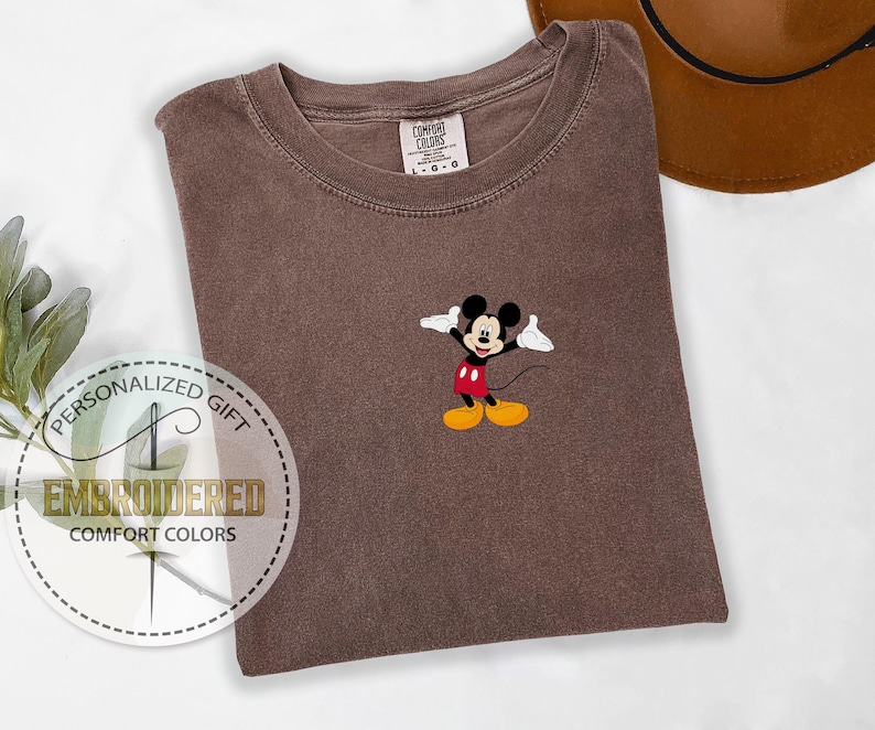 May include: A brown t-shirt with a small embroidered image of Mickey Mouse in the center. The shirt is a unisex style and has a crew neck.