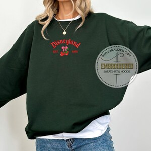 May include: A dark green sweatshirt with the text "Disneyland Est. 1957 1955" and a red and white cherry graphic. The sweatshirt also has a white tag with the text "Personalized Gift Embroidered Sweatshirt & Hoodie".