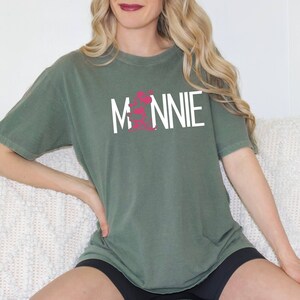 May include: A woman wearing a green t-shirt with the word "MINNIE" printed in white on the front. The shirt has a pink graphic design on the front.