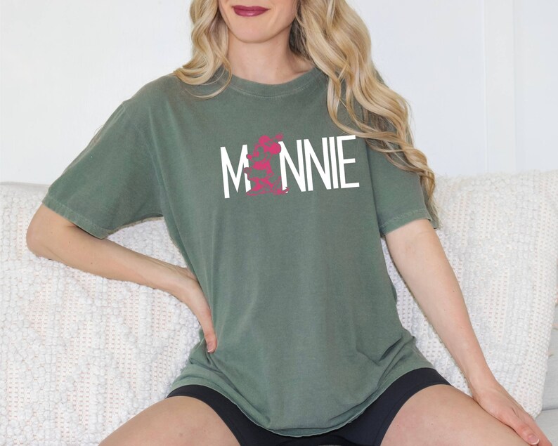 May include: A woman wearing a green t-shirt with the word "MINNIE" printed in white on the front. The shirt has a pink graphic design on the front.