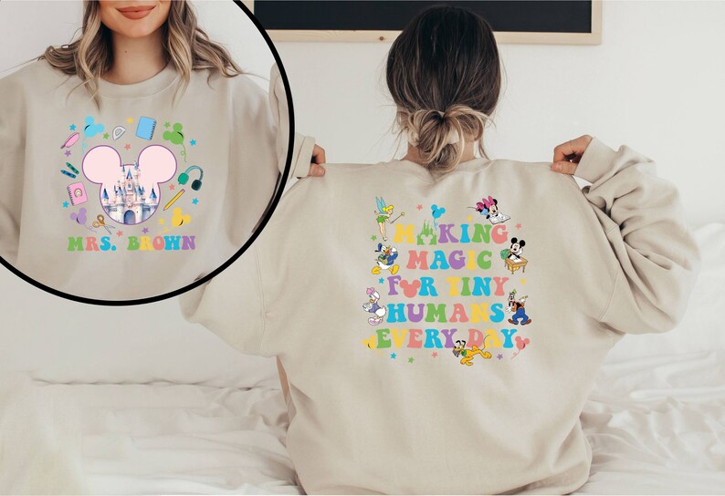 Puede incluir: Sudadera beige con un gr&aacute;fico colorido que presenta una cabeza de Mickey Mouse, un castillo y &uacute;tiles escolares. El texto "MRS. BROWN" est&aacute; debajo del gr&aacute;fico. La parte posterior de la sudadera dice "MAKING MAGIC FOR TINY HUMANS EVERY DAY" con personajes de Disney.