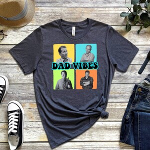 May include: A dark grey t-shirt featuring a graphic of four men in colourful squares. The text "DAD VIBES" is printed in a retro font below the images.
