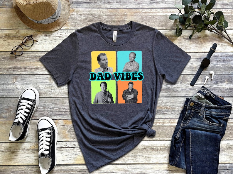 May include: A dark grey t-shirt featuring a graphic of four men in colourful squares. The text "DAD VIBES" is printed in a retro font below the images.