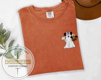 May include: Rust-colored Comfort Colors t-shirt with a Halloween-themed Minnie Mouse ghost design. The graphic features Minnie Mouse in a ghost costume holding a pumpkin. The shirt has a round neckline and a tag that reads "L-G-G".