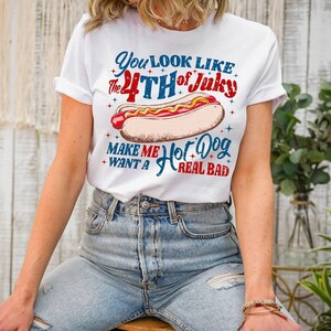 Puede incluir: Una camiseta blanca de manga corta con un gráfico de un perrito caliente con mostaza. El texto dice: You look like the 4th of Juky make me want a hot dog real bad. El diseño utiliza letras rojas y azules con pequeños detalles de estrellas.