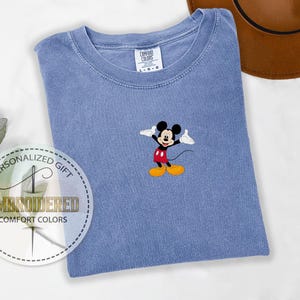 May include: A blue t-shirt with a small embroidered image of Mickey Mouse in the center. The shirt is a light blue color and has a crew neck.