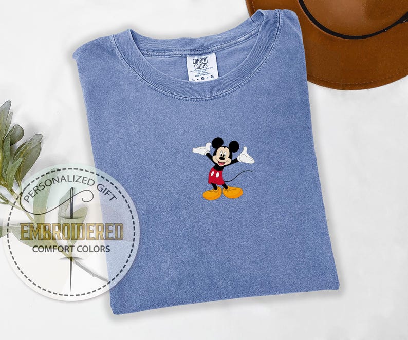 May include: A blue t-shirt with a small embroidered image of Mickey Mouse in the center. The shirt is a light blue color and has a crew neck.