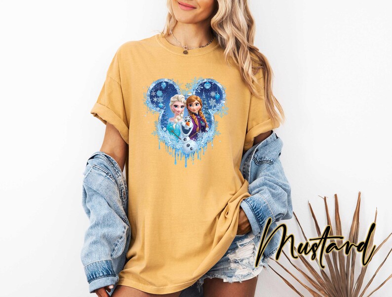 May include: Mustard-colored t-shirt featuring a graphic of Disney's Frozen characters, Elsa, Anna, and Olaf, within a Mickey Mouse-shaped design. The design incorporates blue and white snowflake accents and dripping water effects. The word "Mustard" is written in cursive.