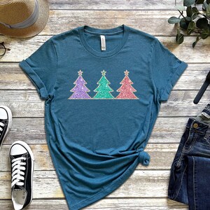 May include: A teal blue t-shirt with three glitter Christmas trees in purple, green, and pink. The trees have gold stars on top.