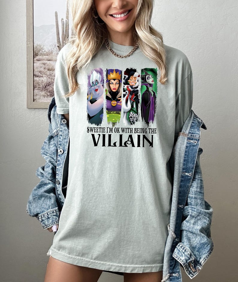 Pu&ograve; includere: T-shirt verde chiaro con quattro personaggi di cattivi dei cartoni animati in pannelli verticali. Il testo "Sweetie I'm OK with being the Villain" &egrave; stampato sotto in nero.