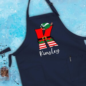 Puede incluir: Delantal de Navidad azul marino con una letra K de elfo roja y verde y el nombre Kinsley impreso en blanco.