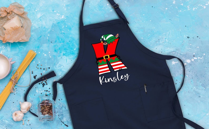 Puede incluir: Delantal de Navidad azul marino con una letra K de elfo roja y verde y el nombre Kinsley impreso en blanco.