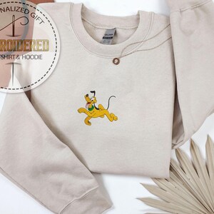 May include: Beige sweatshirt with an embroidered Pluto design. The cartoon dog is yellow with a black tail and ears, and a green collar. The sweatshirt has the text "EMBROIDERED SWEATSHIRT & HOODIE" and "PERSONALIZED GIFT" in a circular design.