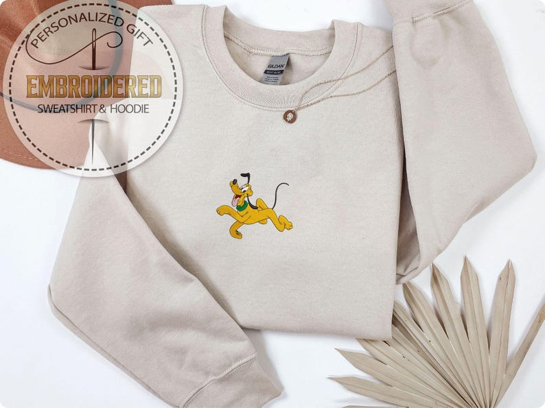 May include: Beige sweatshirt with an embroidered Pluto design. The cartoon dog is yellow with a black tail and ears, and a green collar. The sweatshirt has the text "EMBROIDERED SWEATSHIRT & HOODIE" and "PERSONALIZED GIFT" in a circular design.
