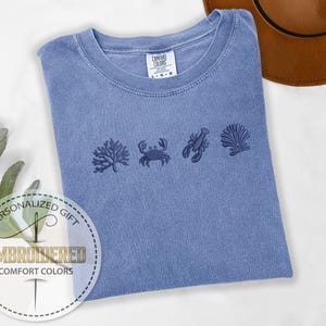 May include: A blue Comfort Colors t-shirt with embroidered sea life designs, including coral, a crab, a lobster, and a seashell. The shirt has a crew neck and a tag that reads "Comfort Colors". A personalized gift label is in the lower left corner.