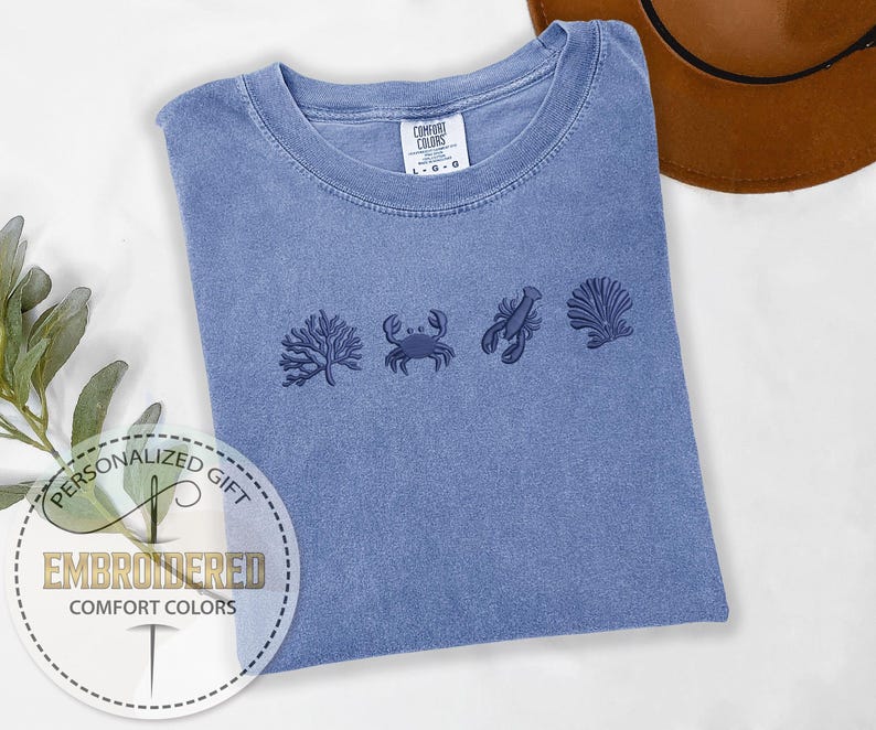 May include: A blue Comfort Colors t-shirt with embroidered sea life designs, including coral, a crab, a lobster, and a seashell. The shirt has a crew neck and a tag that reads "Comfort Colors". A personalized gift label is in the lower left corner.