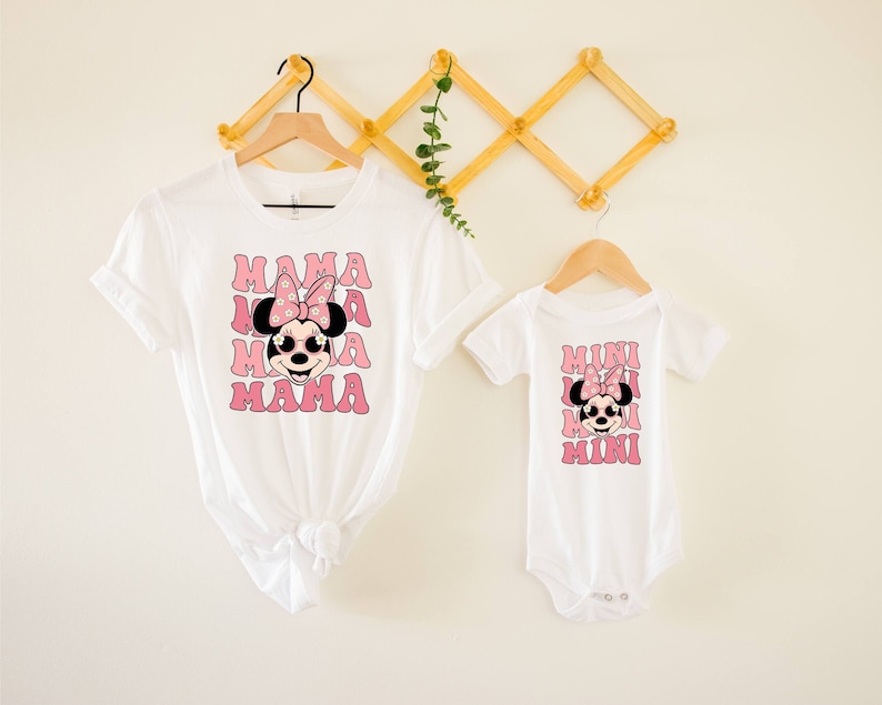 Pu&ograve; includere: Maglietta bianca con una grafica rosa di Minnie Mouse e il testo "MAMA" ripetuto quattro volte. Un body bianco con una grafica rosa di Minnie Mouse e il testo "MINI" ripetuto quattro volte.