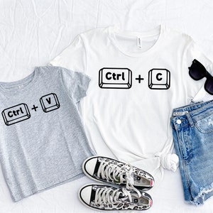 May include: Two gray and white t-shirts with the text "Ctrl + V" and "Ctrl + C" printed on them. The shirts are laid out on a white surface with a pair of leopard print sneakers, a pair of blue denim shorts, and sunglasses.