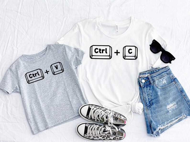 May include: Two gray and white t-shirts with the text "Ctrl + V" and "Ctrl + C" printed on them. The shirts are laid out on a white surface with a pair of leopard print sneakers, a pair of blue denim shorts, and sunglasses.