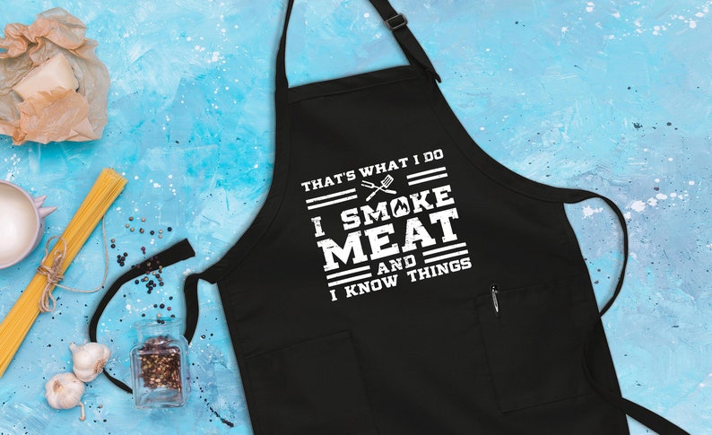 May include: Black apron with white text that reads "That's what I do I smoke meat and I know things". The text is surrounded by white lines. There is a small pocket on the right side of the apron.
