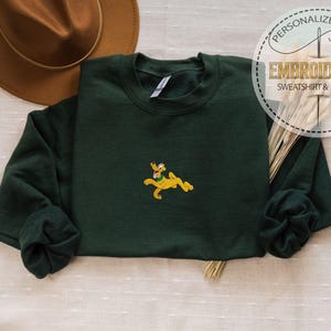 May include: A dark green sweatshirt with a yellow embroidered Pluto design. The sweatshirt has long sleeves and a crew neck. The image also includes a brown hat and text that says "PERSONALIZED GIFT EMBROIDERED SWEATSHIRT & HOODIE".