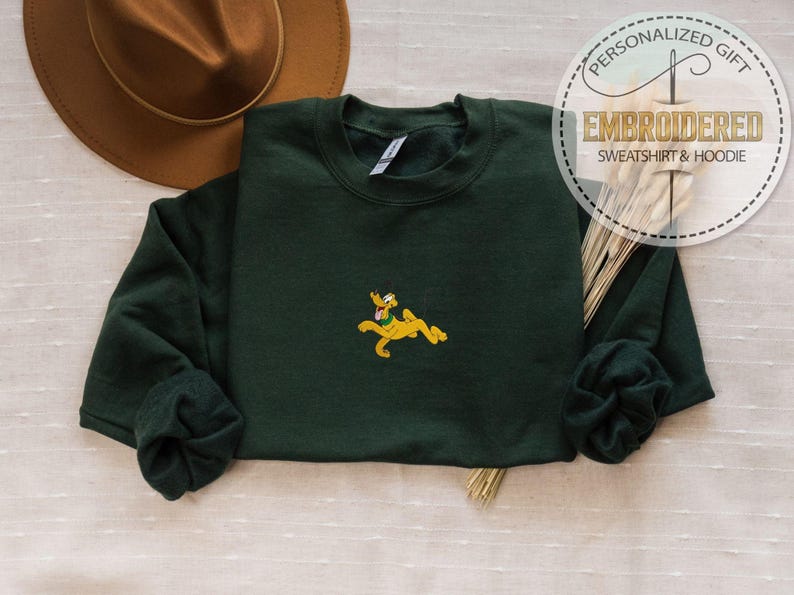 May include: A dark green sweatshirt with a yellow embroidered Pluto design. The sweatshirt has long sleeves and a crew neck. The image also includes a brown hat and text that says "PERSONALIZED GIFT EMBROIDERED SWEATSHIRT & HOODIE".