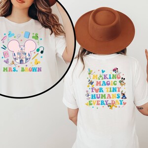Puede incluir: Camiseta blanca con un gr&aacute;fico colorido que presenta un castillo, orejas de Mickey Mouse y &uacute;tiles escolares. La parte delantera dice "MRS. BROWN" y la trasera dice "MAKING MAGIC FOR TINY HUMANS EVERY DAY" con personajes de dibujos animados.