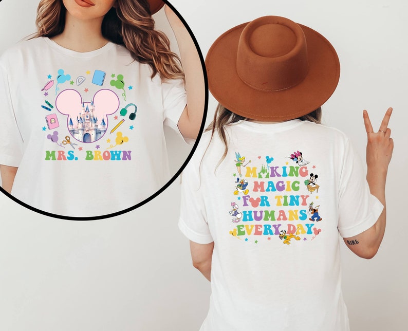 Puede incluir: Camiseta blanca con un gr&aacute;fico colorido que presenta un castillo, orejas de Mickey Mouse y &uacute;tiles escolares. La parte delantera dice "MRS. BROWN" y la trasera dice "MAKING MAGIC FOR TINY HUMANS EVERY DAY" con personajes de dibujos animados.
