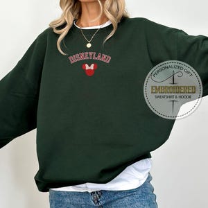 May include: A dark green sweatshirt with the text "DISNEYLAND" in red and white lettering and a red and white Mickey Mouse head with a bow. The text "PERSONALIZED GIFT" and "EMBROIDERED SWEATSHIRT & HOODIE" are in a white circle with a black border.