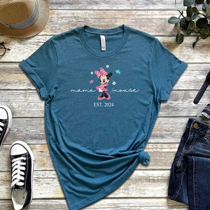 Op de afbeelding: Tealblauw T-shirt met een afbeelding van Minnie Mouse in een roze jurk met witte stippen en een strik. De tekst "mama mouse EST. 2024" is onder de afbeelding gedrukt.
