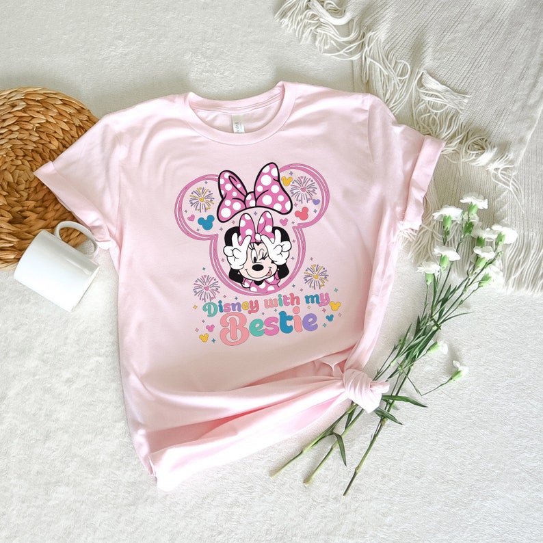 May include: Pink t-shirt with a graphic of Minnie Mouse wearing a pink bow and making peace signs. The text "Disney with my Bestie" is below the image.