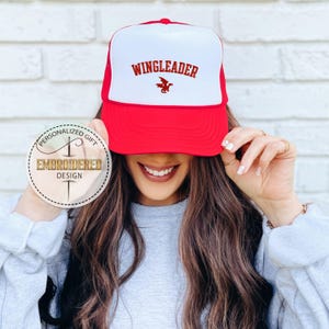May include: A red and white trucker hat with the word "WINGLEADER" in red block letters and a red dragon graphic. The hat has a red brim and a white front panel with a mesh back.