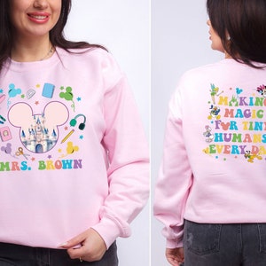 Puede incluir: Sudadera rosa claro con un dise&ntilde;o colorido de Disney. La parte delantera presenta un castillo dentro de un contorno de la cabeza de Mickey, &uacute;tiles escolares y "MRS. BROWN". La parte trasera dice "MAKING MAGIC FOR TINY HUMANS EVERY DAY" con personajes de Disney.