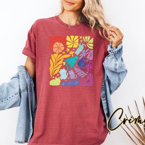 Może przedstawiać: Czerwony t-shirt z kolorowym wzorem kot&oacute;w i kwiat&oacute;w. Grafika przedstawia r&oacute;żne kształty kot&oacute;w w kolorze niebieskim, zielonym, fioletowym i innych, a także kwiaty i gwiazdy. Koszulka ma luźny kr&oacute;j.