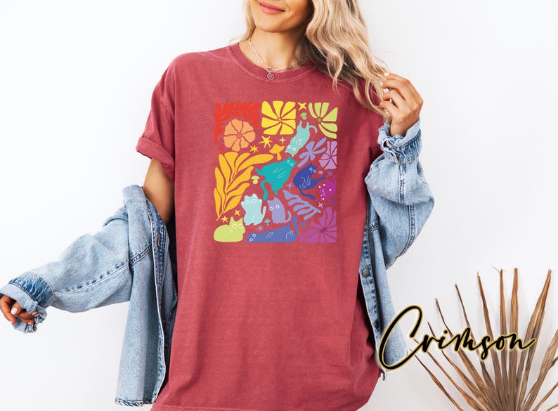 Może przedstawiać: Czerwony t-shirt z kolorowym wzorem kot&oacute;w i kwiat&oacute;w. Grafika przedstawia r&oacute;żne kształty kot&oacute;w w kolorze niebieskim, zielonym, fioletowym i innych, a także kwiaty i gwiazdy. Koszulka ma luźny kr&oacute;j.