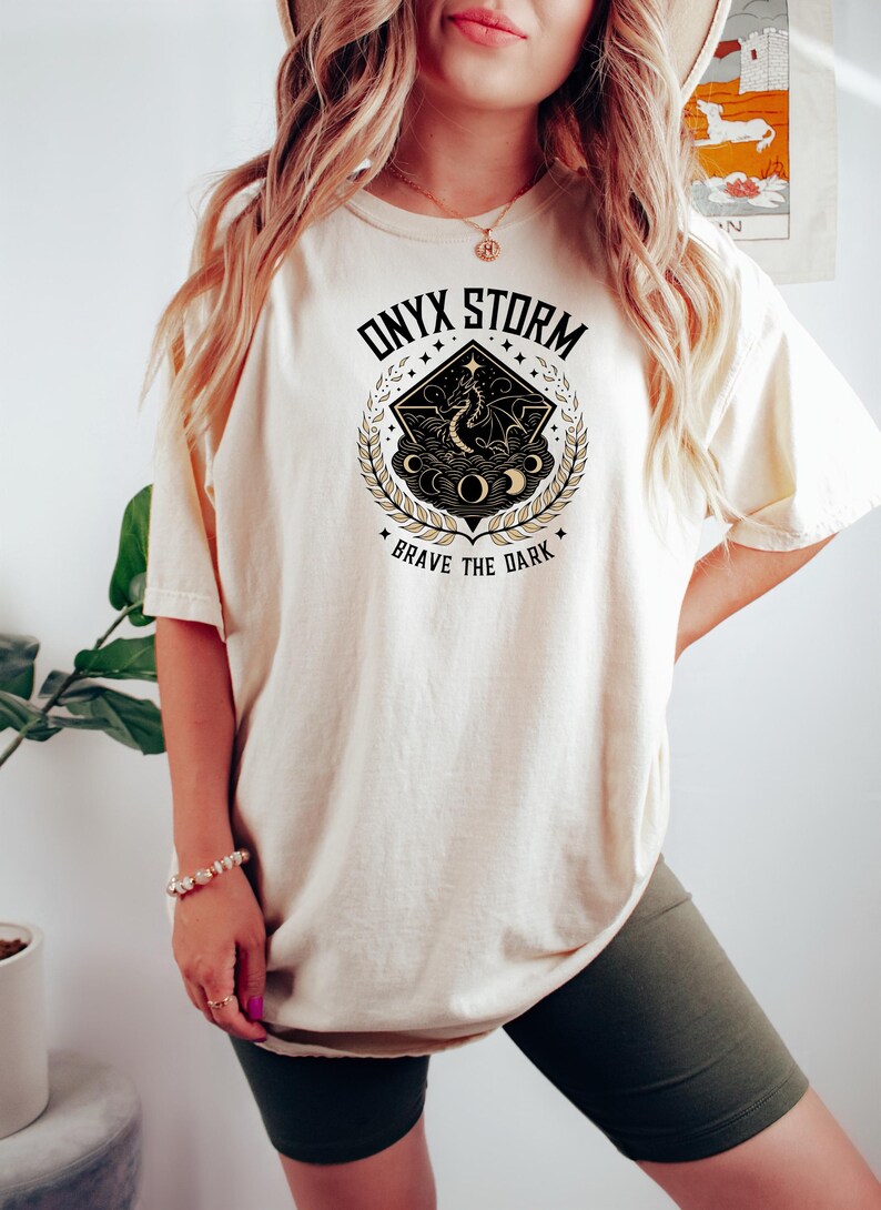 May include: A cream colored t-shirt with a graphic design of a dragon in a circle with the text "ONYX STORM" and "BRAVE THE DARK" in black and gold lettering.