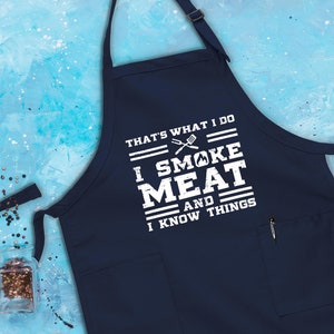 May include: A navy blue apron with a white pocket and the text "That's what I do I smoke meat and I know things".