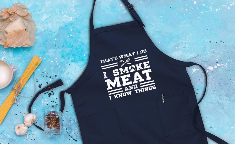 May include: A navy blue apron with a white pocket and the text "That's what I do I smoke meat and I know things".