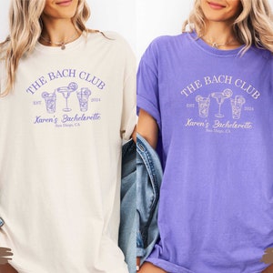 Puede incluir: Dos camisetas con el texto "The Bach Club" y "Karen's Bachelorette" en morado. Las camisetas son de color marfil y violeta.