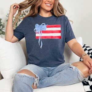 May include: A navy blue t-shirt featuring a watercolour design of an American flag with a blue bow on top. The shirt is being worn by a person sitting on a white sofa.