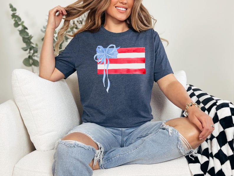 May include: A navy blue t-shirt featuring a watercolour design of an American flag with a blue bow on top. The shirt is being worn by a person sitting on a white sofa.