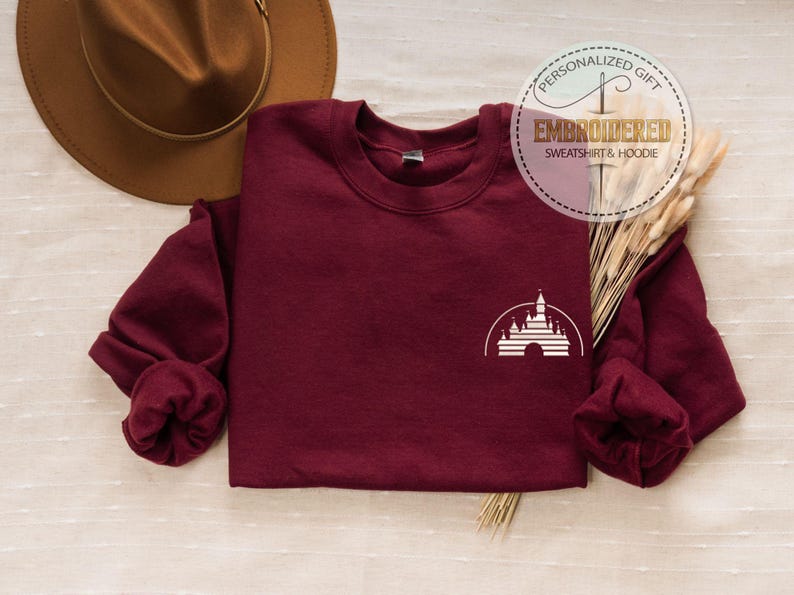 Puede incluir: Sudadera color burdeos con una imagen bordada en blanco del castillo de Disney en la parte delantera. La sudadera est&aacute; doblada y colocada sobre una superficie de color marr&oacute;n claro.