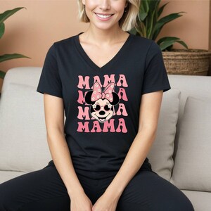Pu&ograve; includere: Maglietta nera a collo a V con una grafica rosa e bianca di Minnie Mouse con occhiali da sole e un fiocco con fiori. Il testo "MAMA" &egrave; ripetuto quattro volte in rosa.