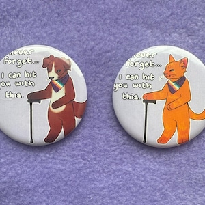 May include: Two round buttons with cartoon animals and text. One button features a brown dog with a rainbow scarf and cane, the other an orange cat. Text reads "Never forget... I can hit you with this."
