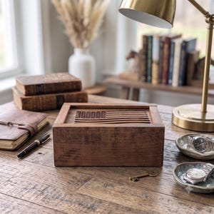 May include: A rectangular wooden box with a carved American flag design on the lid. The box features a dark brown wood grain finish and a smooth, flat surface. It is constructed with visible joinery at the corners and has a solid, sturdy appearance.
