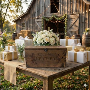 May include: A rustic wooden box with engraved floral details and the names "Jennifer & Wayne KEICHER" sits on a wooden table. White and gold wrapped gifts and a bouquet of white roses and hydrangeas are arranged around the box. A barn is in the background.