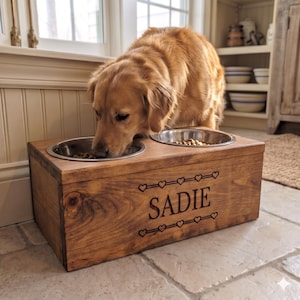 Könnte beinhalten: Ein rechteckiger Futterständer aus Holz mit zwei Edelstahlnäpfen. Auf der Vorderseite ist der Name SADIE in Großbuchstaben eingraviert, flankiert von zwei horizontalen Reihen kleiner Herzsymbole. Ein Golden Retriever frisst aus einem Napf.
