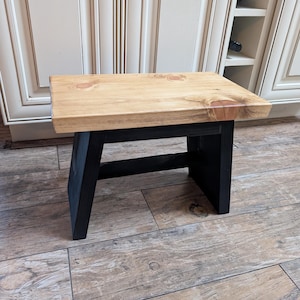 May include: A small wooden stool with a natural wood top and black painted legs. The stool is sitting on a wood floor.