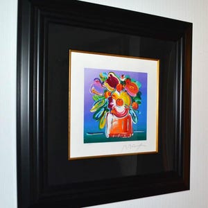 May include: A framed print featuring a vibrant, colorful floral arrangement in a vase against a purple and blue background. The artwork is set within a white mat with a thin gold inner border, enclosed in a wide, black, stepped-profile frame.