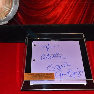 May include: A framed script from "Orange is the New Black" with blue signatures. The script is purple and has the show's title and cast names on a gold plaque. The frame is clear, and the background is red.